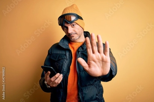 Fototapeta Young handsome skier man wearing ski goggles using smartphone over yellow background with open hand doing stop sign with serious and confident expression, defense gesture