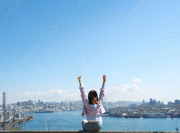 Fototapeta  brunette girl in a purple denim jacket and blue jeans raises her hands up and looks at a large modern sea city with a port and cable-stayed bridge. rear view, freedom of movement, end of quarantine