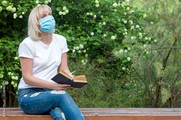 Fototapeta Girl woman in a park on a bench in a medical mask reads a book, walk in the period of quarantine and social isolation, prevention of allergies in the period of seasonal flowering, copy space for text