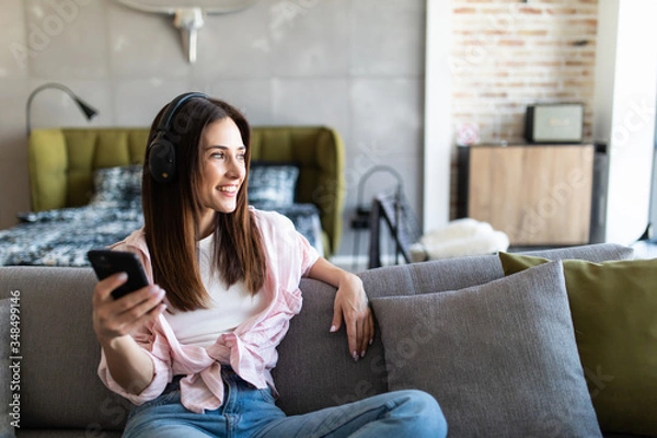 Fototapeta Young woman resting on sofa wearing casual home clothes wireless headphones enjoy weekend free time using online application on cellphone listens music