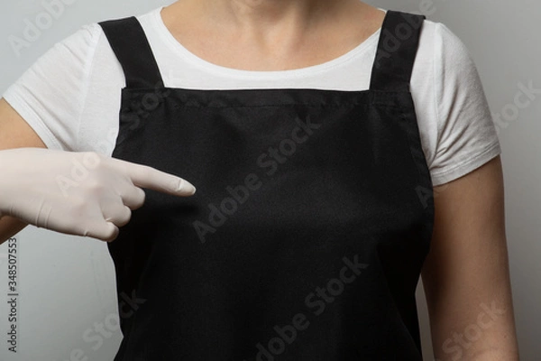 Obraz Closeup, fragment. Girl in black apron pointing finger to empty space on her chest. Protecting gloves. Safety concept. Copy space, place for text, mockup.