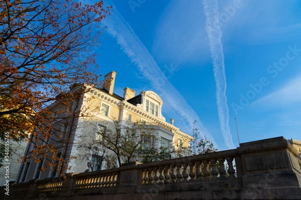 Fototapeta A white mansion pictured in front of the blue sky with two condensation trails, with a tree with orange leaves next to it