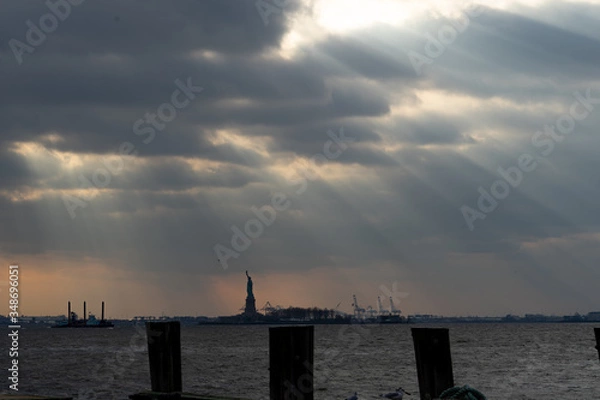 Fototapeta view of the statue of liberty from Manhattan New York on a cloudy afternoon with beautiful sun rays