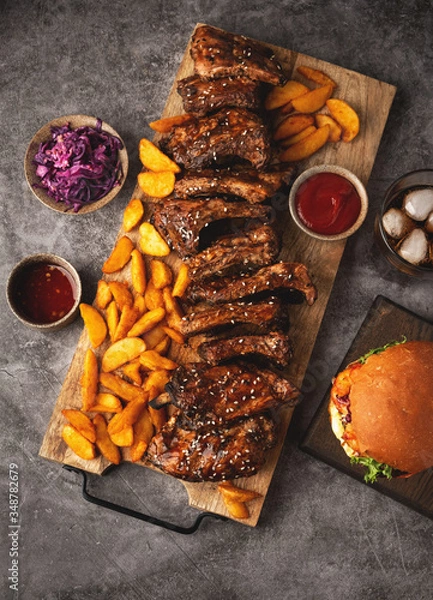 Fototapeta Grilled pork ribs and fried potato wedges on a wooden board, burger and cola glass, sause. Top view, fast food.