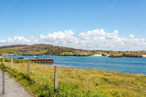 Fototapeta Seafront promenade with wire fence with bay, pier and rocky hills in background, sunny spring day on Inishbofin Island, County Galway, Ireland