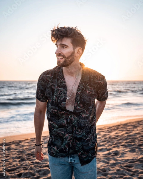 Fototapeta Charming man in casual clothing smiling. Handsome guy standing alone on the beach in sunset and smiling.