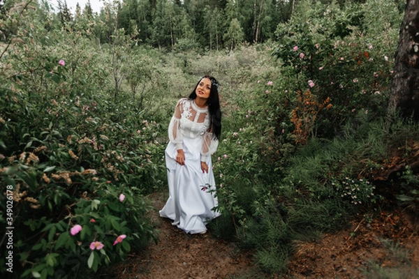 Fototapeta Fairy-tale Princess in a light summer white dress is walking along a wide path towards the wind, a graceful dancing girl in the form of a flower with a fluttering long hem of her dress