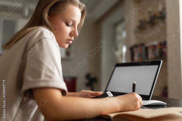 Fototapeta the girl does work from home, checks information from the phone and makes notes in the notebook during quarantine. Home workplace