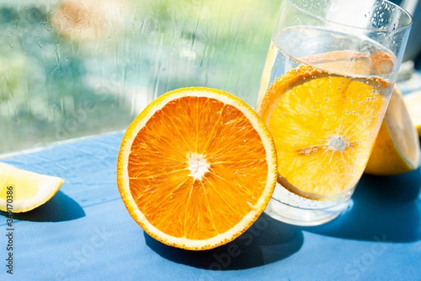Fototapeta half a juicy ripe orange and a glass of transparent water with gases and bubbles and a round slice of orange inside. Take vitamin C, restore health, lead a healthy lifestyle
