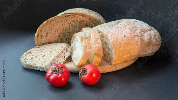 Fototapeta Beautiful bread close-up, two types of bread made from wheat and rye flour and two fresh tomatoes on a round wooden board on a black background. Sliced and half a loaf.