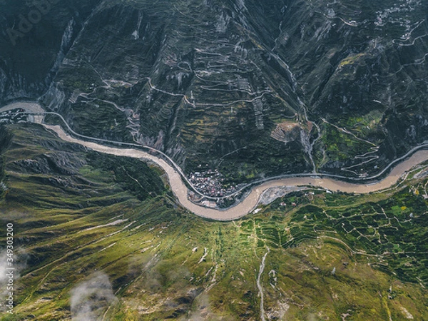 Fototapeta Top down aerial drone view of river and small settlement in between the tall mountains in Tibetan area of Sichuan province, China. Cinematic shot, curved serpentine roads and hills. Journey in spring.
