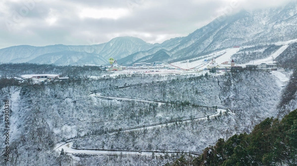 Fototapeta Aerial drone view of Xiling Snow Mountain, the ski resort in Sichuan next to Chengdu. The mountain curved road through the trees, place for skiing and tourist base. Winter sports time.