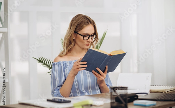 Fototapeta Young girl student sitting at home in the office using book and laptop studing online
