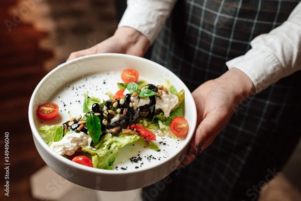 Fototapeta The waiter is holding a white plate with a dish, close-up of hands. Salad with cherry tomatoes, basil and pine nuts