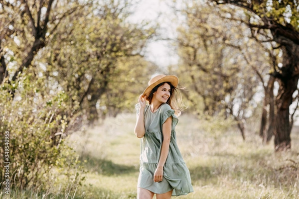 Fototapeta Young woman standing on a tree tunnel background, outdoors in a park, wearing a vintage light green dress and a straw hat.