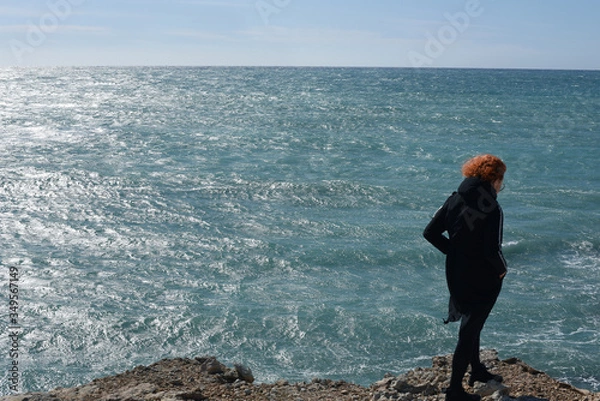 Fototapeta 
A girl with red hair is standing with her back against the background of the sea. The sea sparkles in the sun
