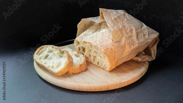 Fototapeta Beautiful bread made from wheat flour on a wooden board on a black background in a paper bag. Sliced slices and half bread.