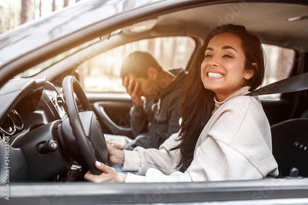 Fototapeta Driving instruction. A young woman learns to drive a car . Her instructor or boyfriend doesn't like the way she drives a car. But the girl is pleased with herself and does not listen to the guy.