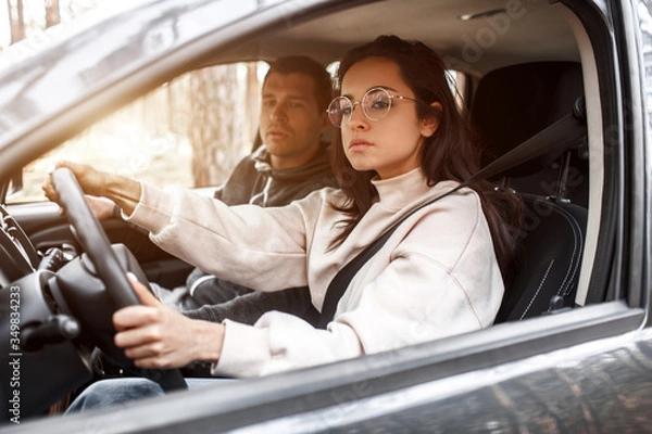 Fototapeta Driving instruction. A young woman learns to drive a car for the first time. Her instructor or boyfriend helps her and teaches her
