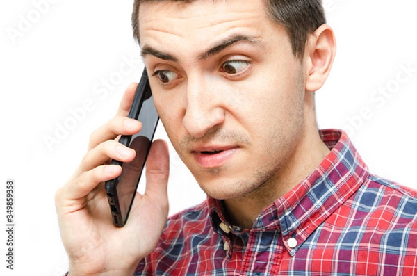Fototapeta Young man in a plaid shirt with a smartphone on a white background. The guy holds a smartphone by the ear, talking on the phone. Anger, anger, surprise, attentiveness during a telephone  conversation