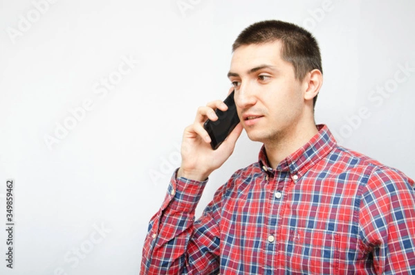Fototapeta Young man in a plaid shirt with a smartphone on a white background. The guy holds a smartphone by the ear, talking on the phone. Anger, anger, surprise, attentiveness during a telephone  conversation
