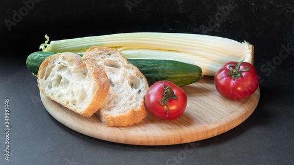 Fototapeta Two slices of white beautiful airy white bread, fresh vegetables - cucumbers, tomatoes and celery, all on a round wooden cutting board on a black background close-up.