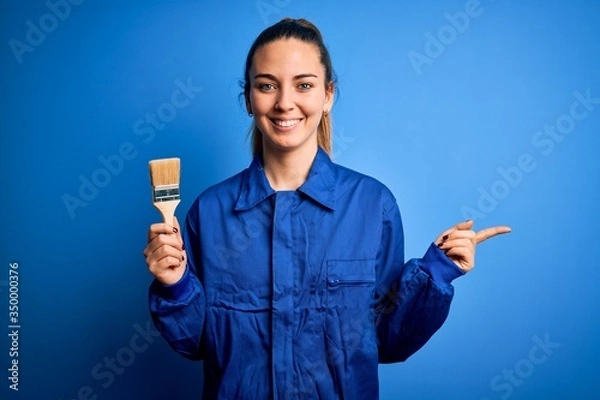 Obraz Young beautiful painter woman with blue eyes painting wearing uniform using paint brush very happy pointing with hand and finger to the side