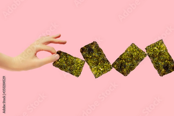 Fototapeta Female hand hold crispy seaweed on pink background with copy space. Dry nori sheets korean or japanese roasted food texture. Flat lay.