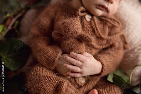 Obraz A newborn charming boy, 5 days old, sleeps in a cozy bear outfit and in a light brown outfit. He sleeps in a basket and holds a tiny teddy bear.