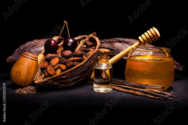 Fototapeta Bottle of oil with cocoa pod and cocoa beans, cherry, bourbon vanilla pods, tobacco pipe and glass jar of honey. Still life on a black background.