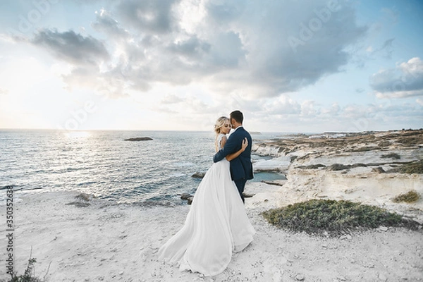 Obraz Newlywed couple hugging together on the white rocks at the seacoast at Cyprus. The bride in a wedding dress the groom in a suit posing with beautiful landscape on the background
