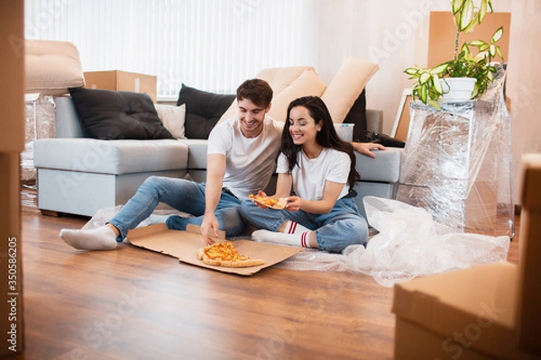 Fototapeta Happy family eating pizza on moving day. Picture of a young couple enjoying rest time while sitting together in the new house.