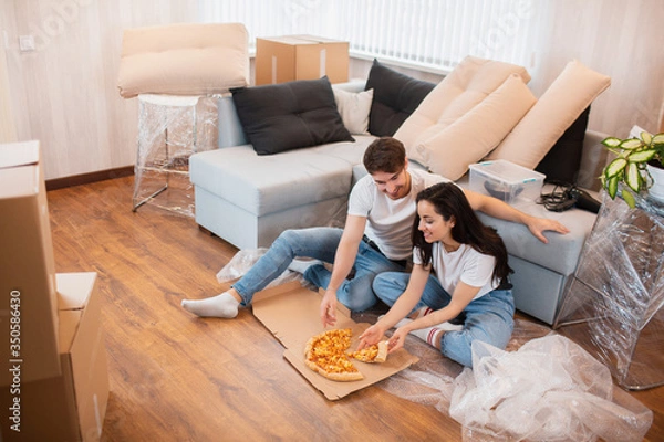 Fototapeta Happy family eating pizza on moving day. Picture of a young couple enjoying rest time while sitting together in the new house.