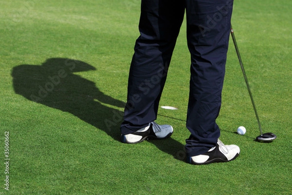 Fototapeta A man with shadow is standing on the green of a golf course.