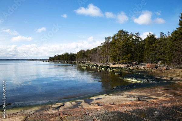 Fototapeta A coastline along the smooth sea.  Large jagged rocks, boulders and cliff in the foreground. The sky is pale blue and cloudy. 
