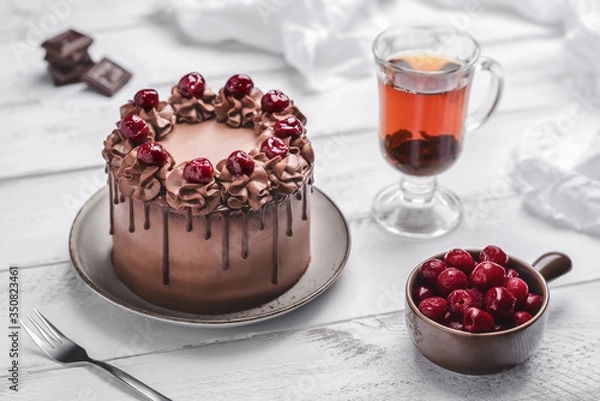 Fototapeta Cherry chocolate cake ornate with cream, cherries and dripping chocolate on grey plate, glass teacup, small bowl with cherries, chocolate bar pieces, napkin and fork on white wooden background