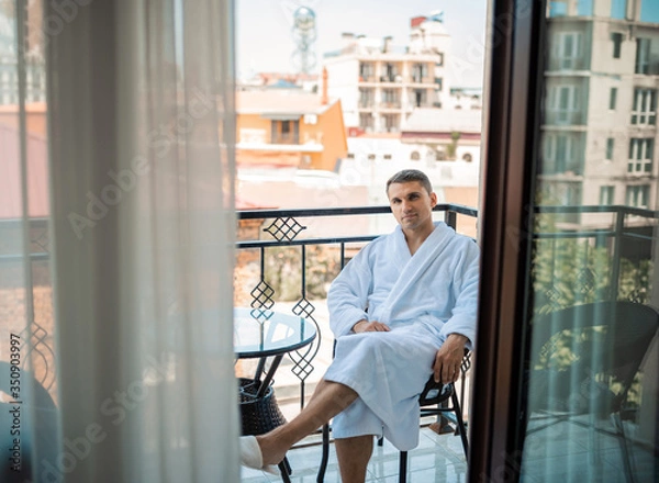 Fototapeta Handsome bearded man in the morning sitting in a hotel room in a robe. Young man wearing white bathrobe after shower bath. Gygiene concept