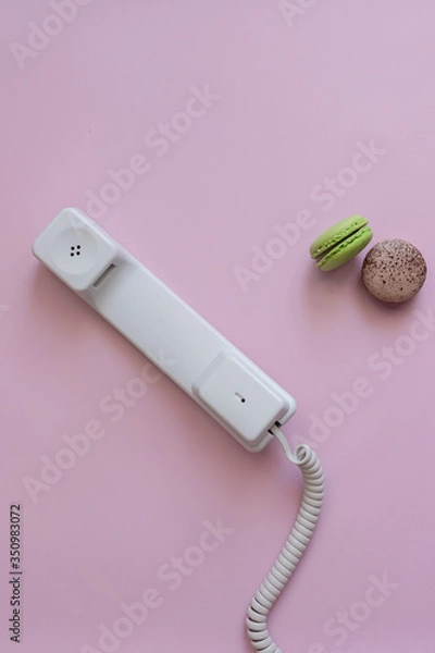 Fototapeta A close up view of A white vintage dial telephone handset and two macaronis on pink background. Communication device. A retro receiver with phone curly wire and copy space. Call center. Office context