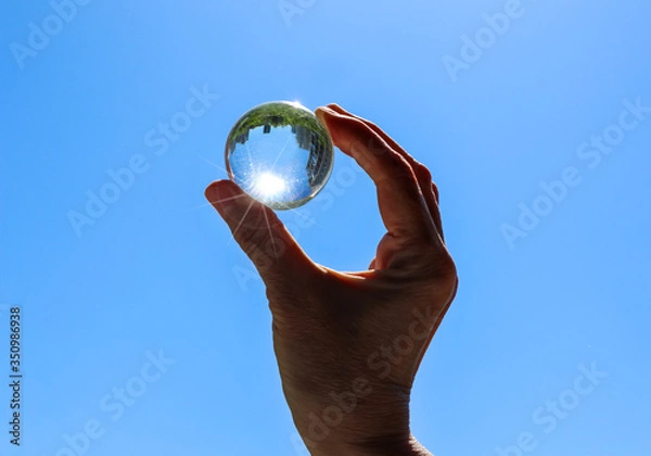 Obraz Female hand holding a transparent crystal ball with city and trees reflection. Round glass sphere with sunbeams against a blue sky background. Bright summer view.