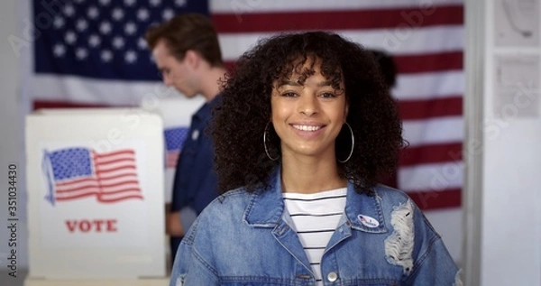 Fototapeta MCU Młoda Latynoska w dżinsowej kurtce z nową naklejką „Głosowałem”, uśmiechnięta i stojąca przed wyborcami w lokalach wyborczych z flagą USA w tle