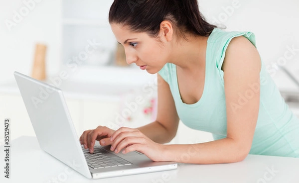 Fototapeta Young woman at her laptop in kitchen