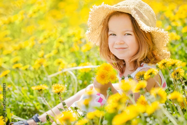 Fototapeta little cute girl 3 years old smiling in a straw hat sits on the grass in yellow dandelions in the summer with a place for text