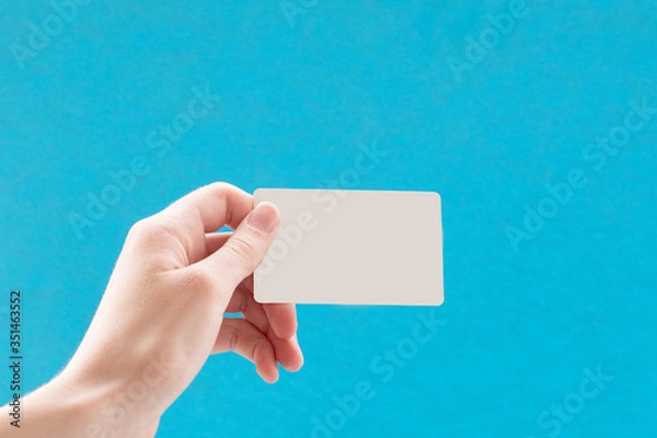 Fototapeta Close up of a female hand holding a blank white business card on a blue background. Business idea, copy space