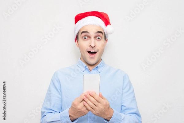 Fototapeta Amazed guy in Santa Claus hat with phone, portrait, white background. Concept of a new year.