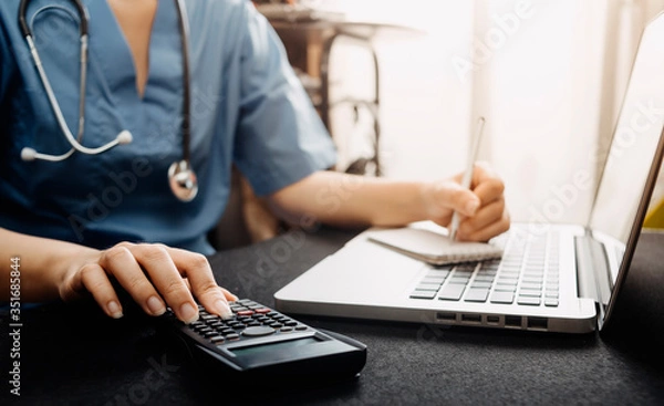Fototapeta Healthcare costs and fees concept.Hand of smart doctor used a calculator and smartphone, tablet for medical costs at hospital in morning light