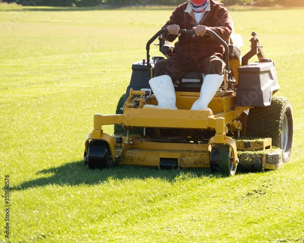 Fototapeta Soft focus of Gardener use Lawn mover on green grass in the modern garden, Machine for cutting lawns, the orange sunlight splashes behind and the green background fence, mow the lawn service concept.
