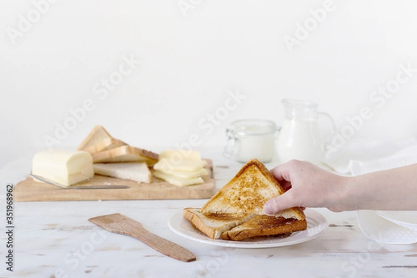 Fototapeta Hand takes fried croutons from the table. In the background - butter, cheese, milk, sour cream. Wooden white background. Country style. Breakfast.