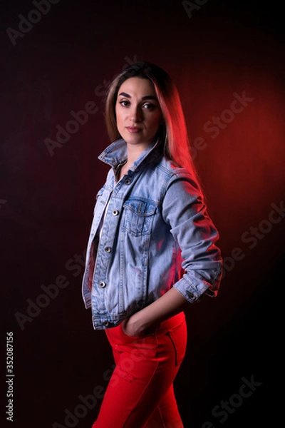 Fototapeta A young girl in a denim jacket and red trousers on a black background with red lighting
