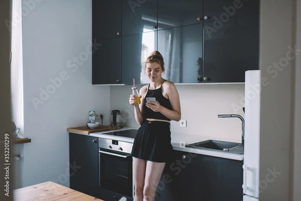 Fototapeta Cheerful female drinking fresh beverage and browsing mobile phone