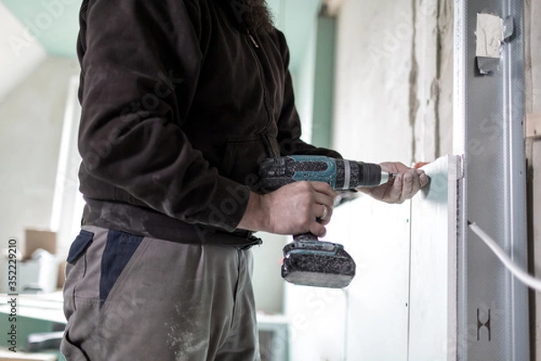 Fototapeta Ein Handwerker befestigt eine Gipsplatte mit einer Bohrmaschine an der Wand